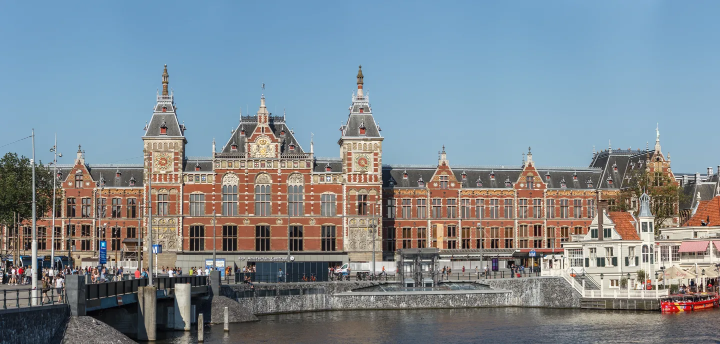 De historische bakstenen gevel en torens van Amsterdam Centraal met water en kade op de voorgrond