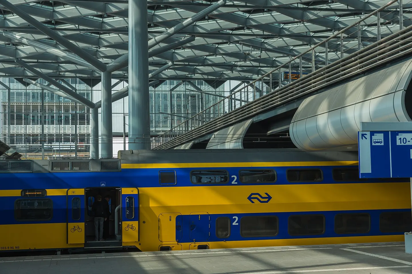 Een geel-blauwe dubbeldekker van NS langs het perron onder de stalen overkapping van Den Haag Centraal