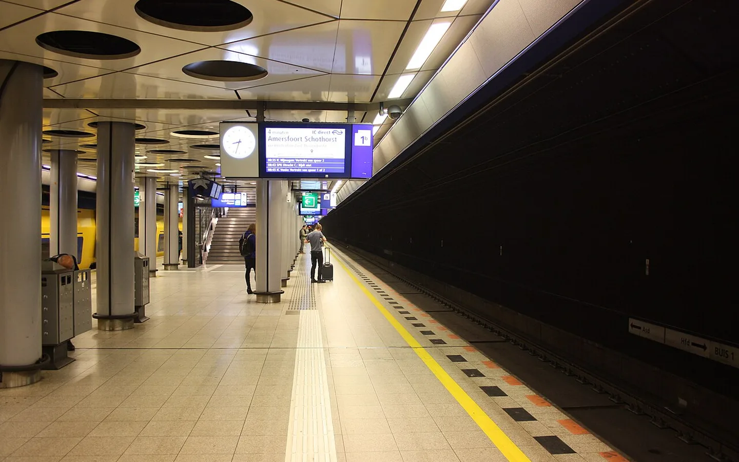 Het ondergrondse perron van station Schiphol Airport met zuilen, vertrekbord en een reiziger met koffer